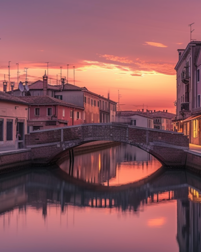 Sunset over Comacchio, Italy's canals, showing reflective water with pastel-colored houses and the iconic Trepponti Bridge in the background, locals walking along cobblestone paths.