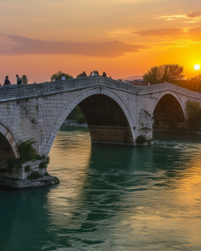 Warm sunset hues bathe the historic Stone Bridge and Vardar River in Skopje, with locals and travelers enjoying the picturesque scene.