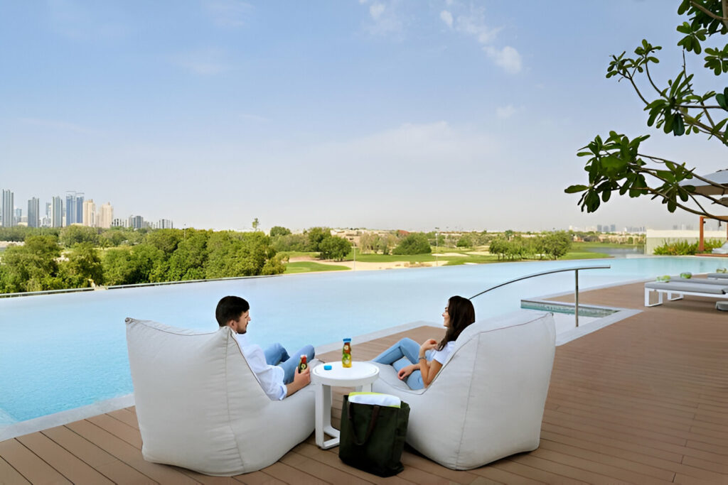 Two individuals sitting on white cushions by a pool of a hotel in Dubai